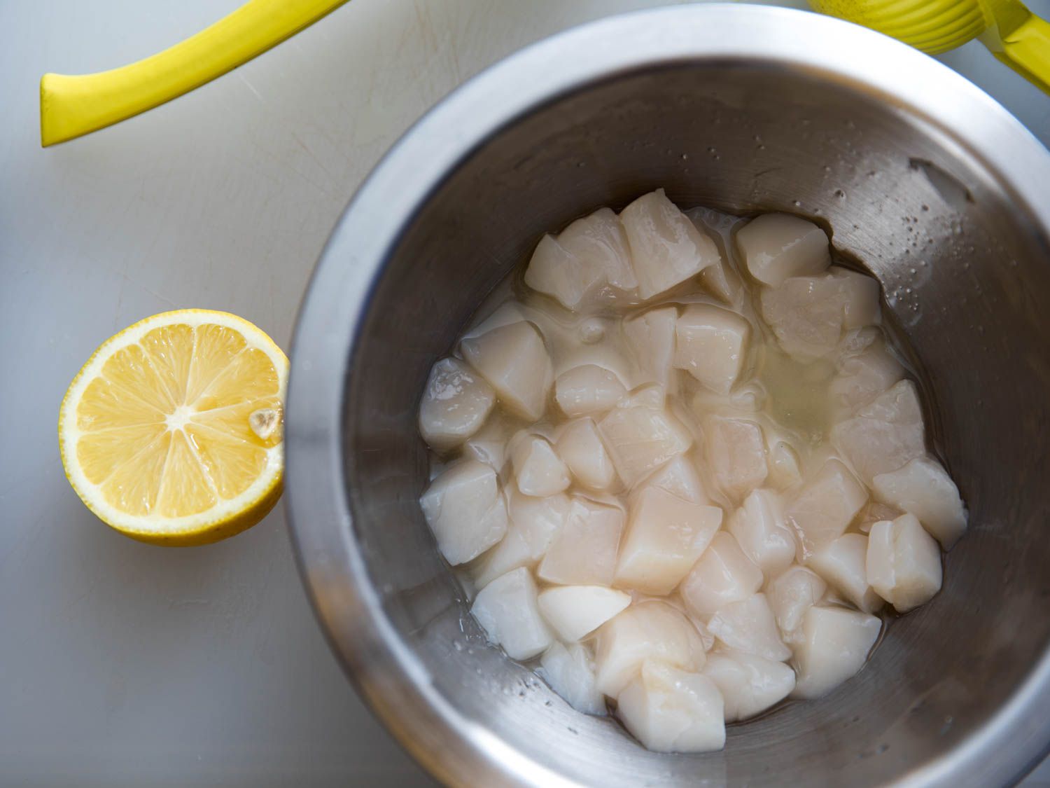 生扇贝浸泡在装满柠檬汁的不锈钢碗里，旁边放半个柠檬和黄色榨汁机。gydF4y2Ba