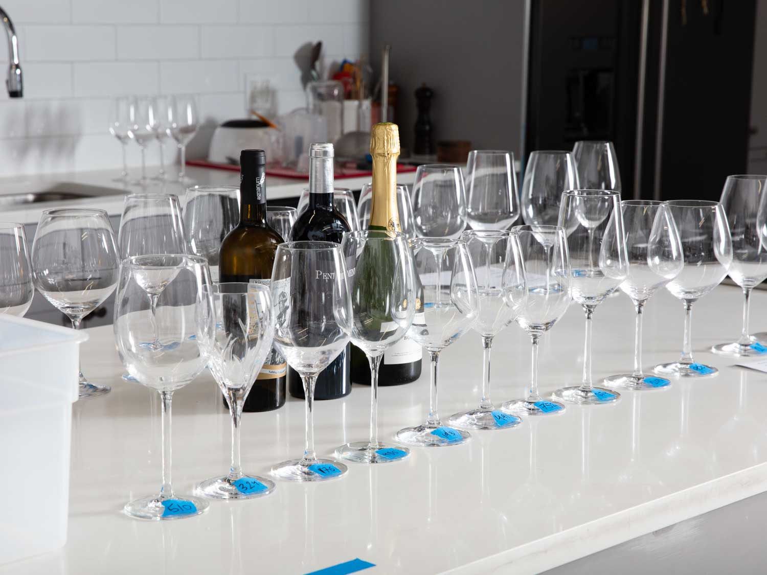 工作台面上排列着几排通用的酒杯，底座上有蓝色的标签，旁边是红色、白色和起泡酒的瓶子