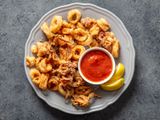 A round gray plate with a pile of fried calamari, two lemon wedges, and a small bowl of marinara sauce.