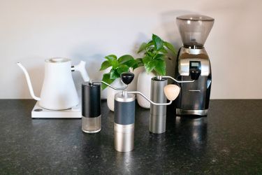 three handheld grinders are arranged on a counter in front of a kettle and electric grinder