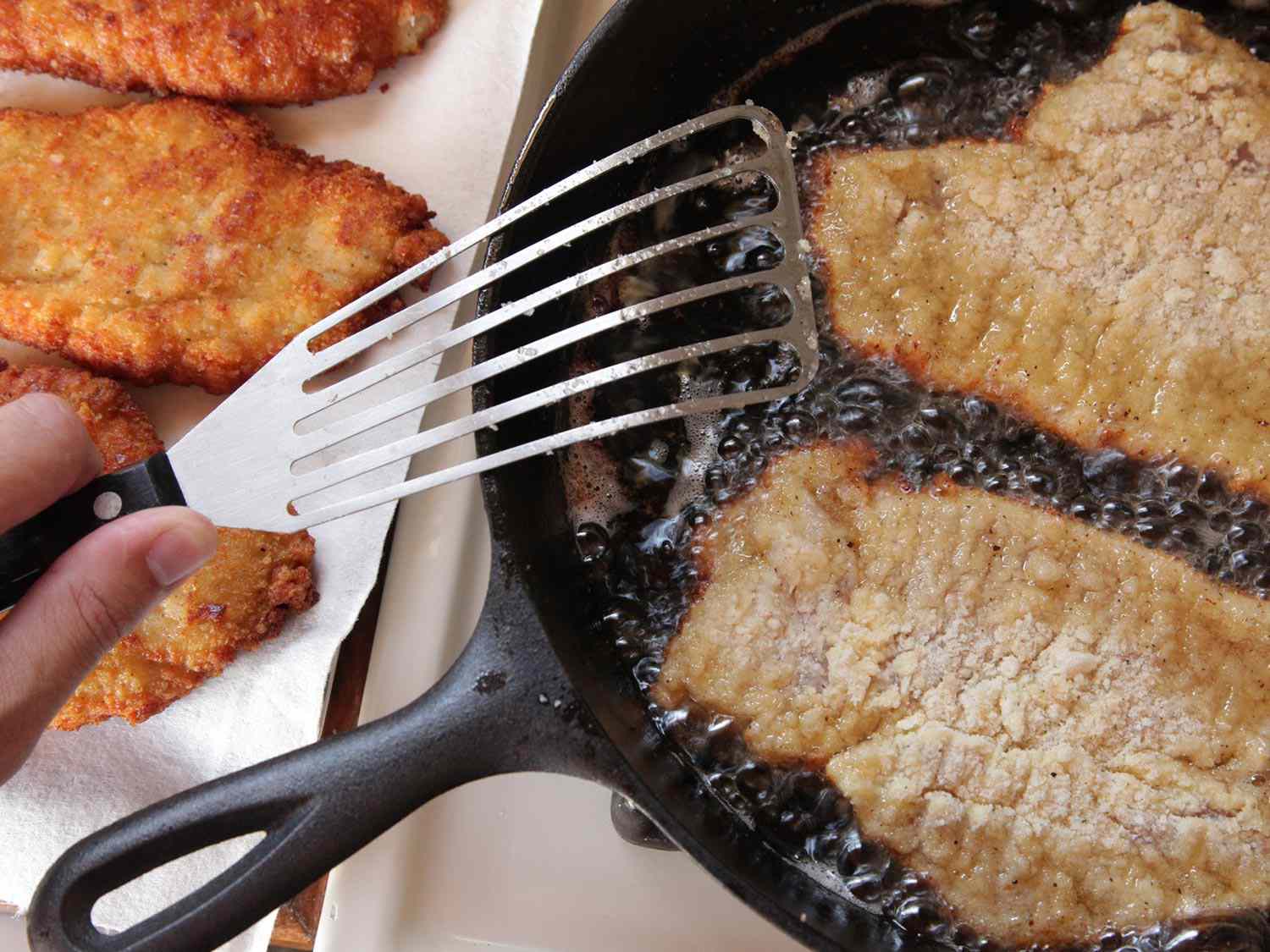 Finished fried chicken breasts laid on paper towel beside more chicken breasts deep-frying in cast iron skillet