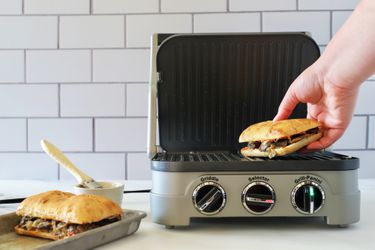 a person placing a sandwich onto a panini press and a small sheet tray holding another sandwich sitting besides the panini press