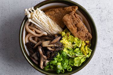 Japanese udon noodles in a ceramic bowl with mushroom-soy broth, stir-fried mushrooms, raw mushrooms, sliced scallions, cabbage, and tofu.