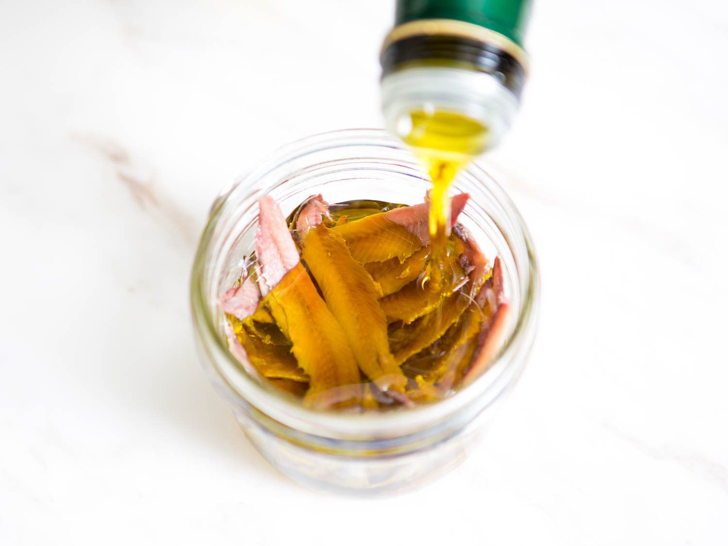 Drizzling olive oil over anchovy fillets in a glass bowl.