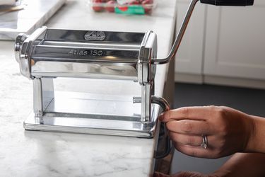 Two hands adjusting the clamp of a manual pasta maker on the side of a countertop