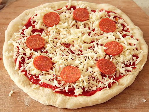将披萨面团做成圆形，浇上酱汁、奶酪丝和意大利辣香肠，放在木质披萨皮上，准备进入烤箱。