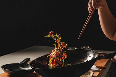 在黑暗的厨房里，一个人拿着筷子扔锅，导致五颜六色的食物悬浮在空中