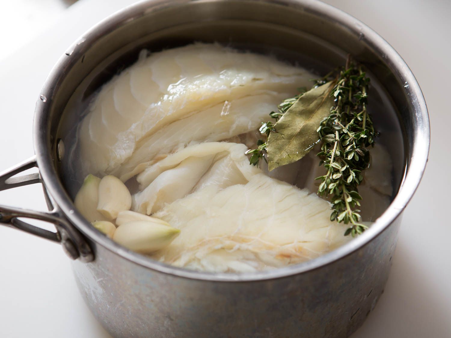 A stainless steel pot containing salt cod, water, garlic, fresh thyme, and a dried bay leaf.