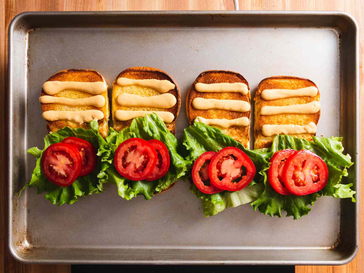 烤盘上放着四个烤好的汉堡面包，每个面包的上半部分涂上三行自制的Shack酱，下半部分涂上生菜和番茄。