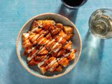 Overhead of a small serving bowl of patatas bravas drizzled with salsa brava and allioli, with glasses of red and white wine on the side.