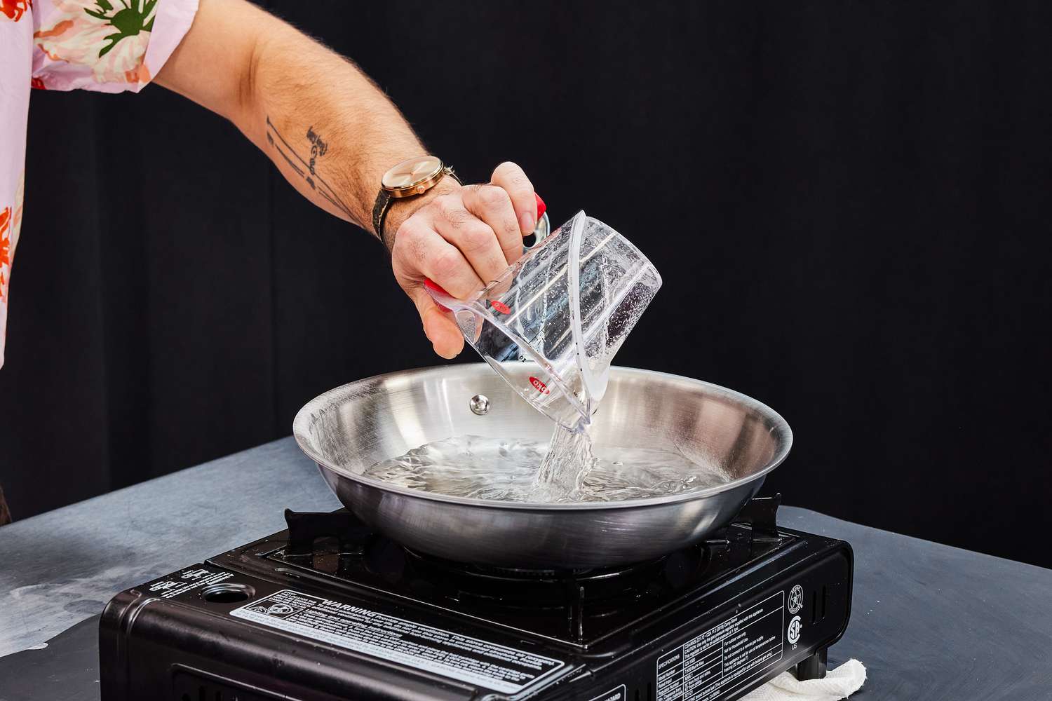 a person pouring water from a measuring cup into a stainless steel skillet