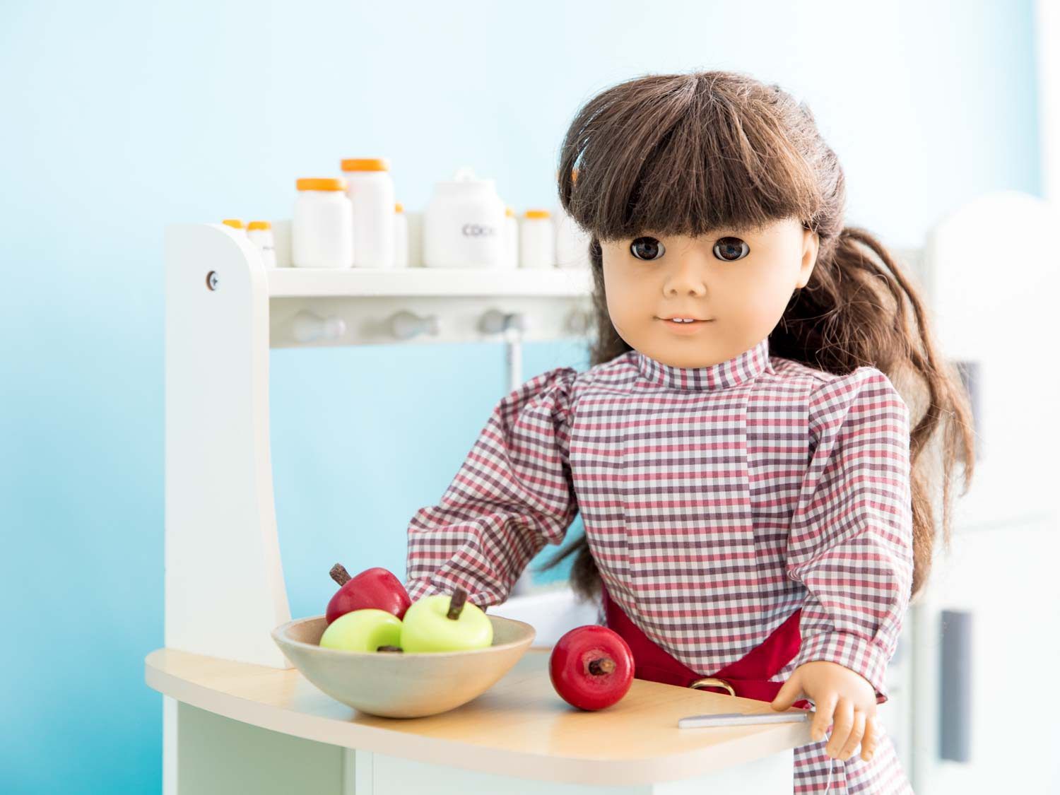 在一个白色的娃娃厨房里，一个萨曼莎娃娃站在柜台前的一碗苹果旁
