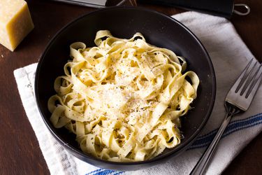 A black ceramic bowl of cooked homemade fettucine noodles sprinkled with black pepper and parmesan cheese.