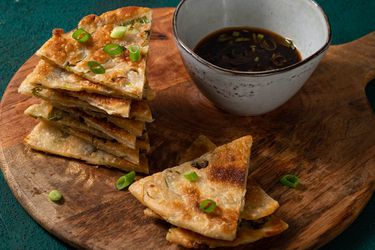 Scallion pancakes cut into slices, stacked on top of each other on a round wooden cutting board with a ceramic bowl of dipping sauce.