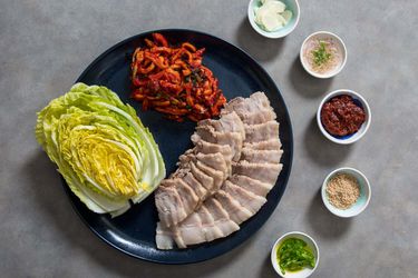 A platter of bossam, complete with sliced pork belly, Fresh cabbage leaves, and an array of condiments.