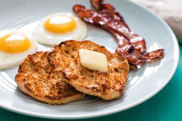 把自制的英式松饼撒上黄油，放在蓝色盘子上，旁边是两个单面鸡蛋和两片培根。