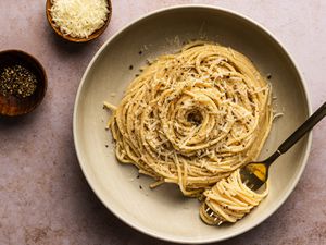 Cacio e pepe on a tan ceramic plate alongside two small bowls, one holding shredded cheese and the other holding freshly ground pepper. There is a metal fork with pasta swirled around it on the plate.