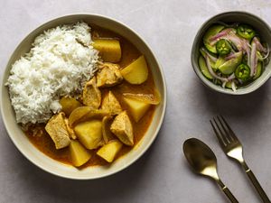 一碗泰式咖喱鸡，放在奶油色的陶瓷碗里。图片右侧是一个小碗，里面盛着黄瓜沙拉。在图片的右下角有一把叉子和勺子。