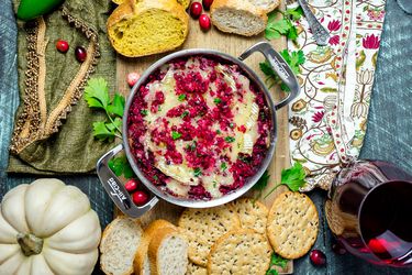 Cranberry Jalapeno Baked Brie on a cutting board with sliced bread and crackers. A glass of red wine is next to the cutting board.
