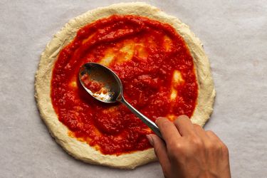 New York-style pizza sauce being spread on an uncooked pizza dough round with a metal spoon.