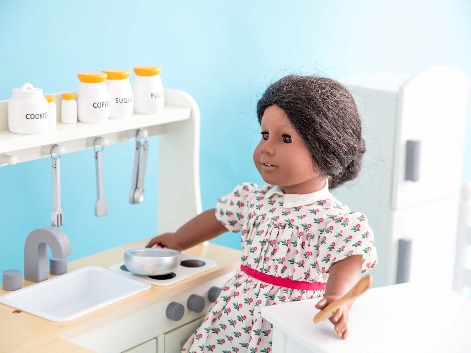 一个阿迪娃娃站在白色的娃娃厨房里，拿着炉子上的煎锅和木勺