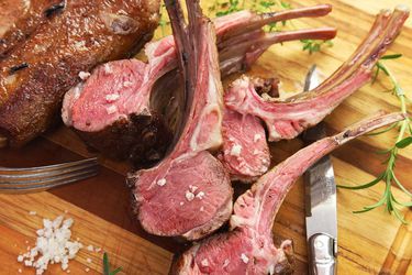 A rack of lamb cooked sous vide and sliced into individual portions, all fanned out on a wooden cutting board with a pile of salt and sprigs of fresh herbs all around.