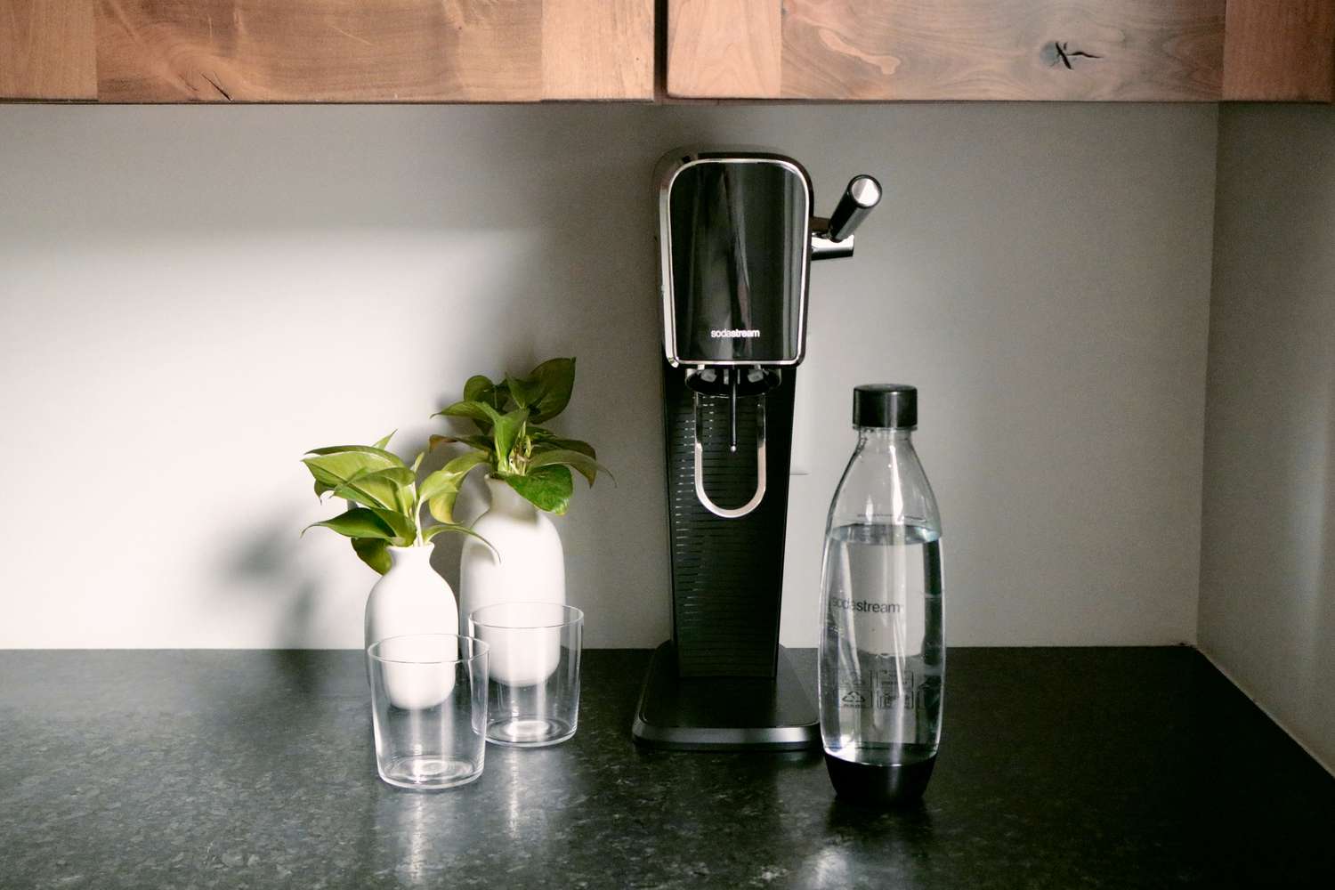 A black SodaStream on a kitchen countertop with two glasses and a SodaStream bottle beside it