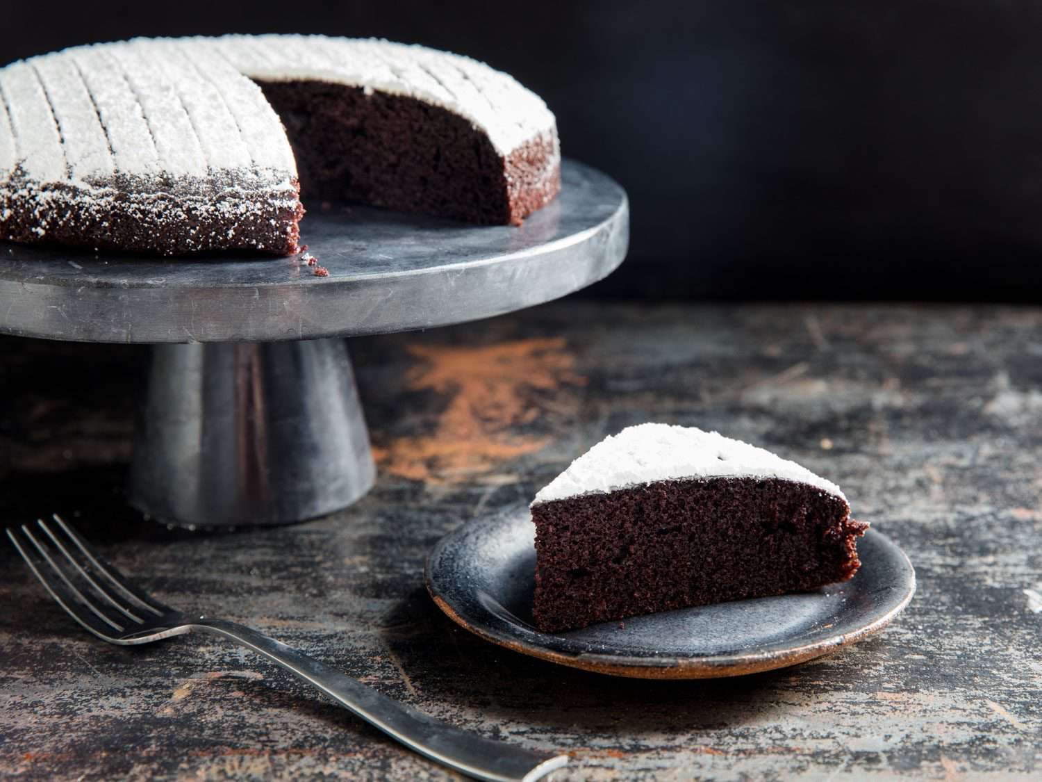 一块巧克力橄榄油蛋糕放在盘子里，旁边是蛋糕架上的完整蛋糕