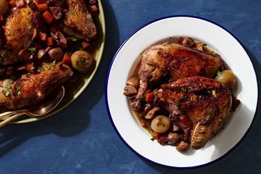 A white, blue-rimmed plate holding a large serving of Coq au Vin. On the lefthand side of the image is a round platter holding more of the Coq au Vin.