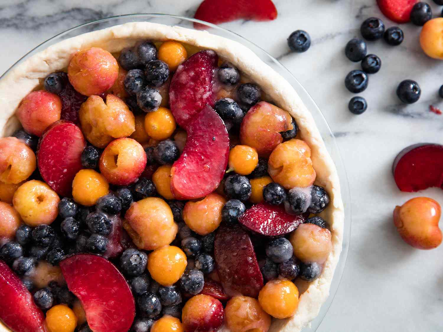 开销的蛋糕盘在一个白色的大理石板。蛋糕盘内衬面团,充满了蓝莓、醋栗角,雷尼尔山樱桃,李子片。水果已经被扔进sugar-starch混合物和额外的水果是分散盘的一边。gydF4y2Ba