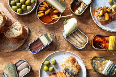 overhead shot of an array of tinned fish, or conservas, on a wooden table with bread and olives.