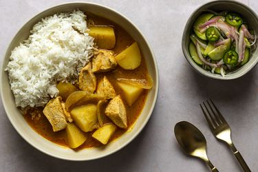 泰国咖喱鸡黄色的一碗奶油颜色的陶瓷碗。图像的右边是一个小碗里附带的黄瓜沙拉。有一个叉子和勺子在图片的右下角。