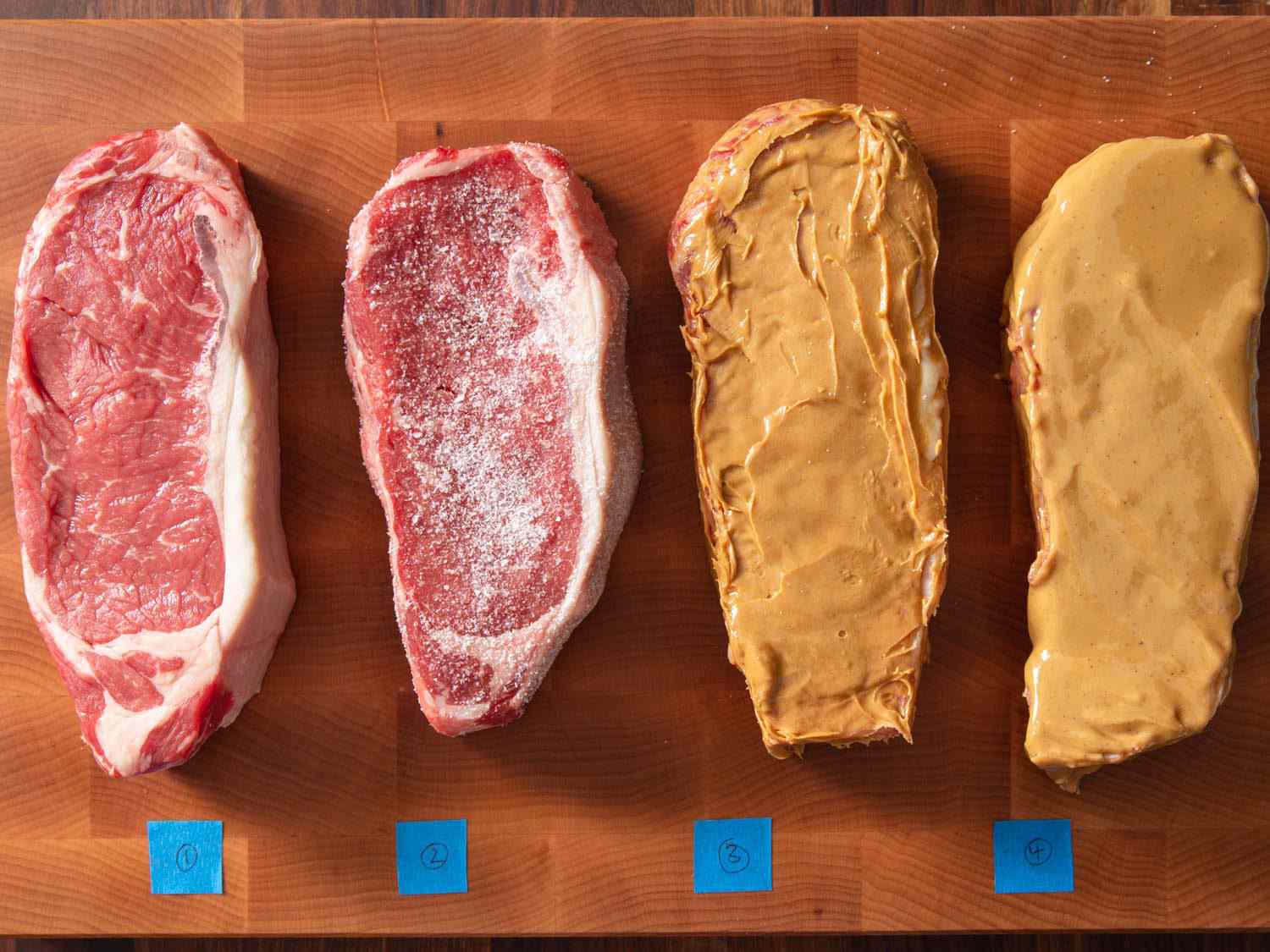 在砧板上由数字胶带标识的四条带牛排的俯视图。从左到右:普通牛排;大量加盐的牛排;涂有Skippy花生酱的牛排;牛排上再涂一层天然花生酱。