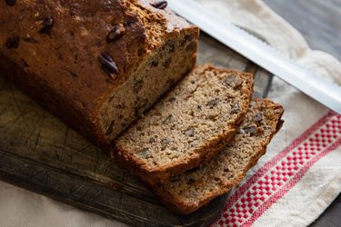 Banana bread with pecans sliced on a small wooden cutting board on top of a white kitchen towel with knife on the side.