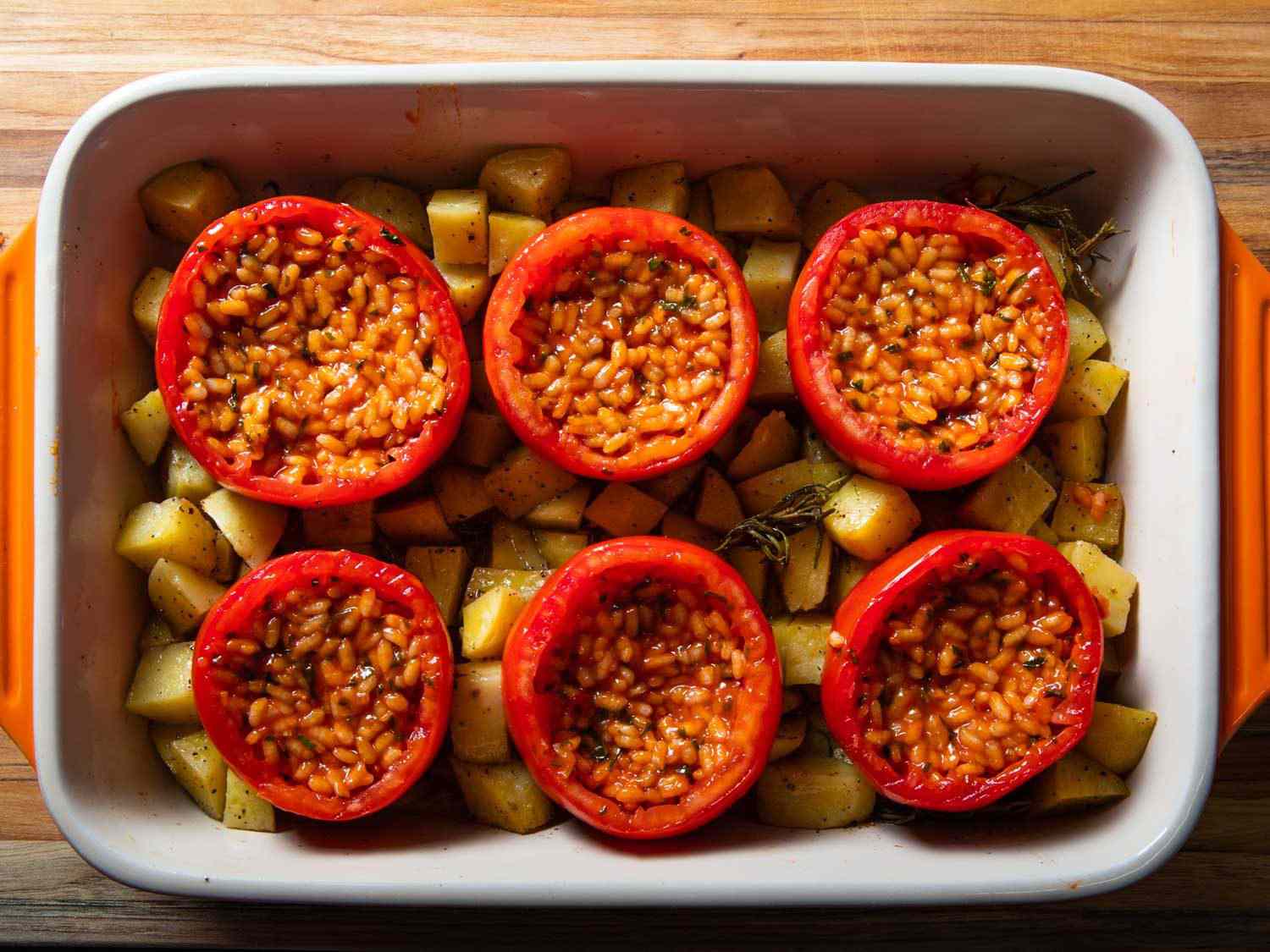 Overhead of rice-stuffed tomatoes in a baking dish with potatoes before being roasted.