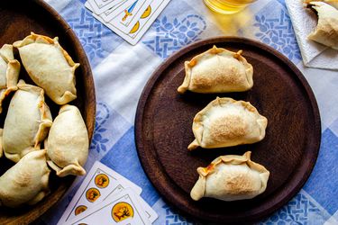 Overhead view of a table scene. Two plates of empanadas are placed on top of a traditional Argentinian fabric with a blue and white checkered. In the middle of the frame is a dark wooden table with 3 empanadas in a line, surrounded by a beer, playing cards and a half eaten empanada.