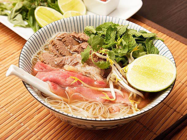 A bowl of pho, garnished with herbs, beef slices and lime wedges.