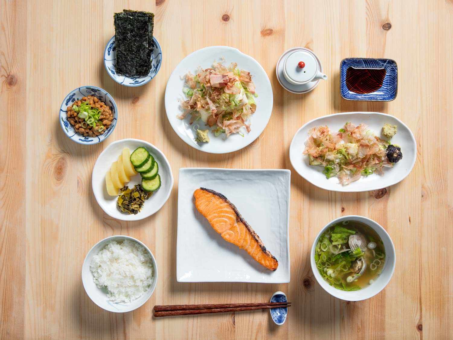 俯拍日式早餐:米饭、泡菜盘、纳豆、海苔、豆腐、酱油瓶、酱油、茄子、味增汤、木桌上的咸鱼。