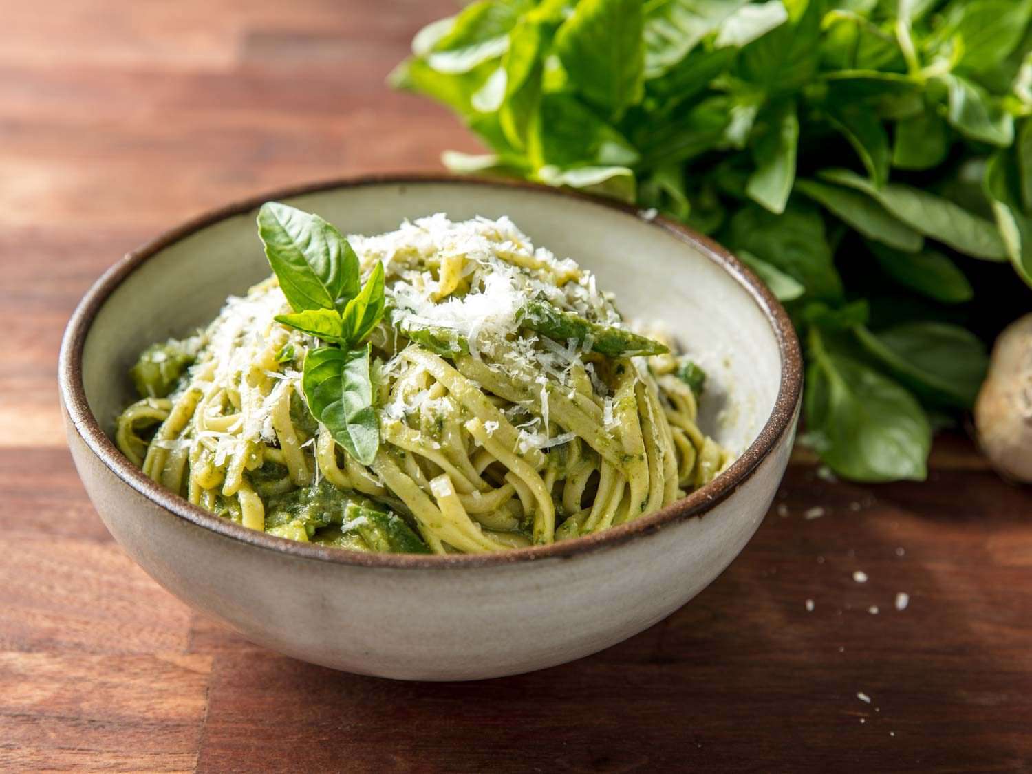 Pesto on linguine in a stone bowl, topped with cheese and basil leaves, with basil leaves in the background.