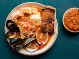 A serving of cioppino in a bowl, overflowing with plump shrimp, mussels, clams, calamari, fish, and more. There's a piece of deeply toasted sourdough on the side, and a small bowl holding a roasted red pepper condiment.