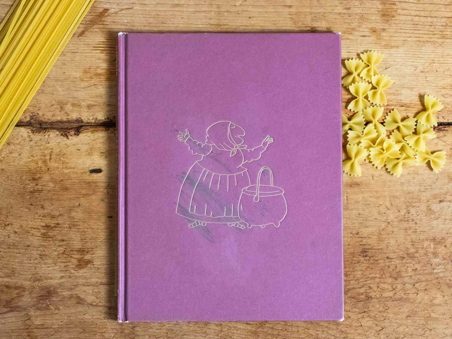 A copy of the book Strega Nona by Tomie dePaola, on a wood tabletop with dried pasta. .