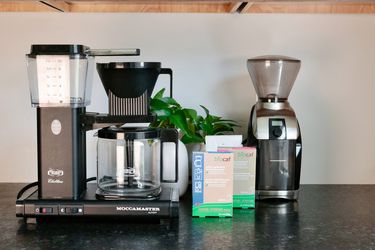 A coffee maker sitting next to two packages of descaler and cleaner and a burr grinder