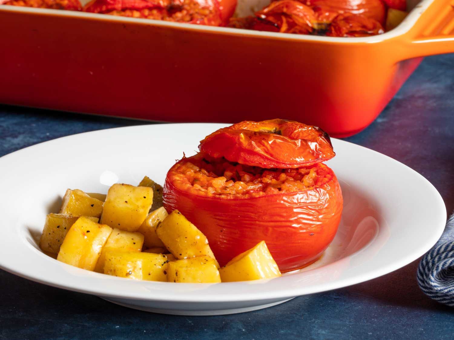 Close-up side view of a plated rice-stuffed tomato with roast potatoes on the side.