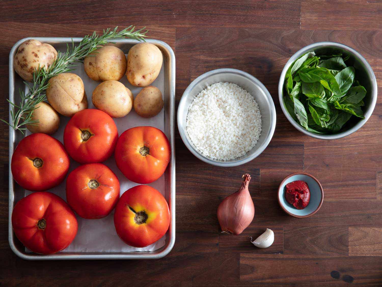 Overhead of ingredients for rice-stuffed tomatoes: potatoes, fresh rosemary, tomatoes, arborio rice, shallot, garlic, tomato paste, and fresh basil.