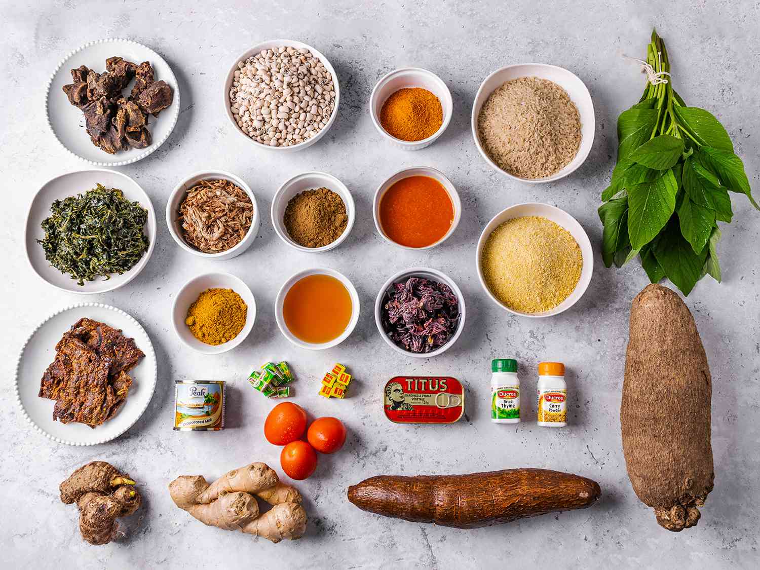 Overhead view of all the essential ingredients needed in a pantry to cook a range of Nigerian dishes. Ingredients range from spices to roots to tinned food and fresh spices, organized in a neat grid