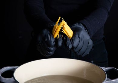 一双黑色手套的手打破干面条面条一半在一个搪瓷铸铁壶开水。