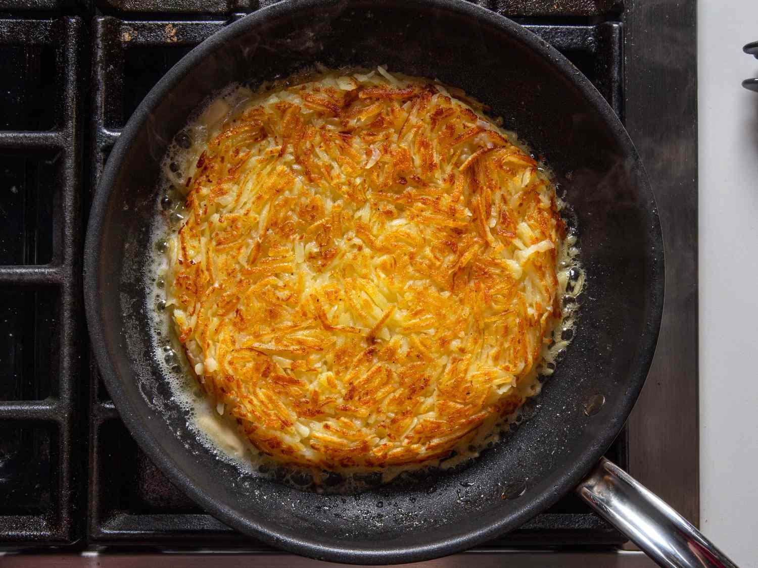 Cooking the rÃ¶sti in the skillet until the second side is browned and crisped. Foaming butter can be seen around its periphery.