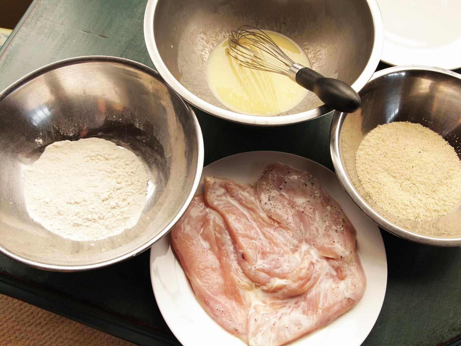 Breading set-up for chicken parmesan: a plate of skinless, boneless chicken breasts, a bowl of flour, a bowl of egg wash, and a bowl of breadcrumbs.