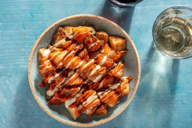 Overhead of a small serving bowl of patatas bravas drizzled with salsa brava and allioli, with glasses of red and white wine on the side.