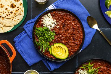 A large bowl of vegetarian bean chili topped with sliced avocado, scallions, cilantro, and white onion. The periphery of the image contains a wide variety of plates and bowls holding tortillas, sliced limes, and additional chili.